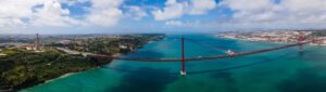 A panoramic view of the 25 de Abril Bridge spans the Tagus River in Lisbon, a must-see holiday destination in Europe. The red bridge, akin to the Golden Gate, links the city to Almada. Blue skies with scattered clouds hover above as lush greenery and urban areas line the riverbanks.